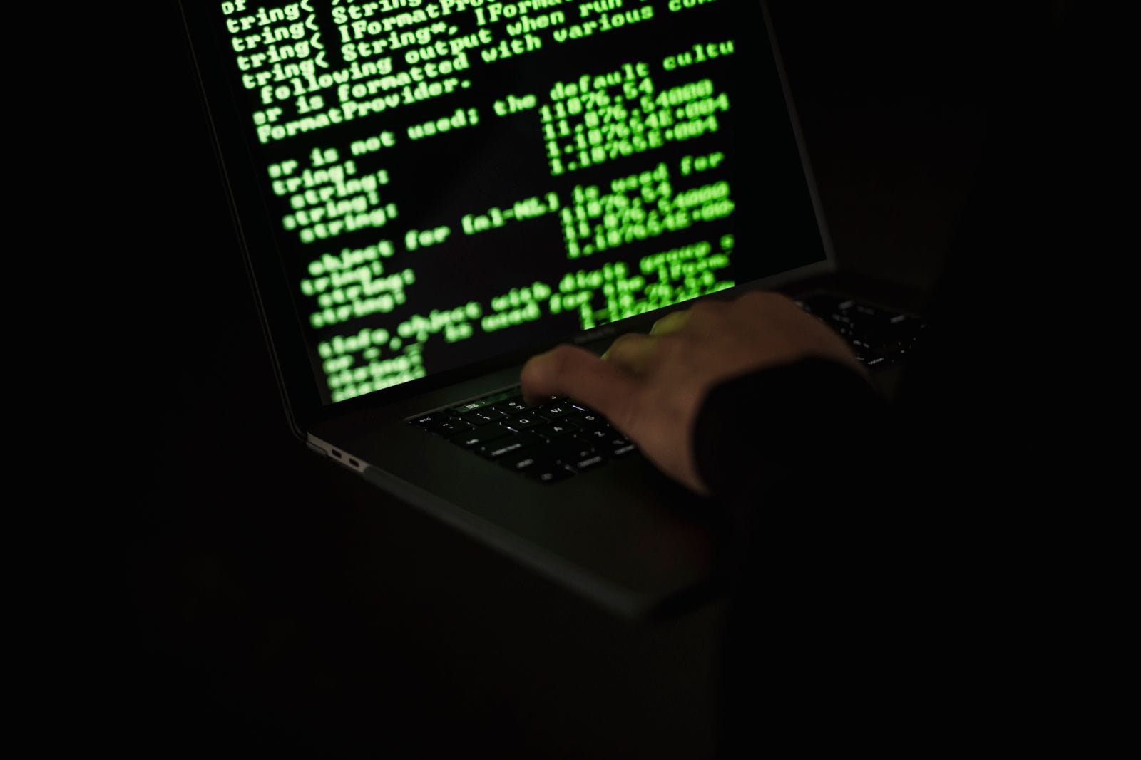 A person typing at a laptop displaying green terminal code in a dark room