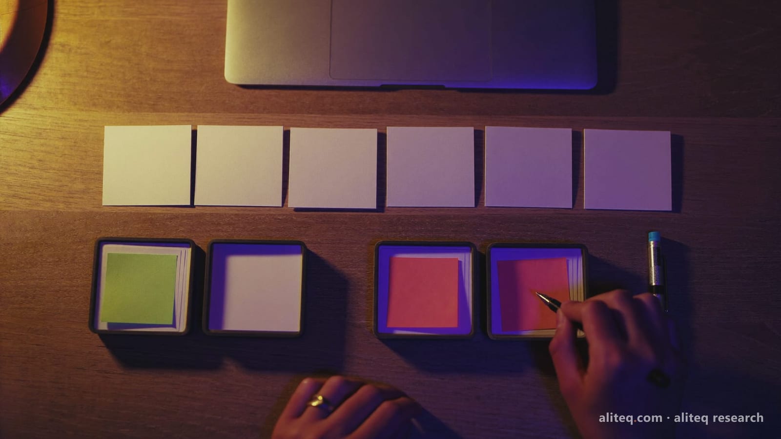 Overhead photo of a desk at night with a row of blank white cards above four small trays, one tray holding green sticky notes and another coral ones, while a hand with a pen sorts cards, lit in warm and violet light