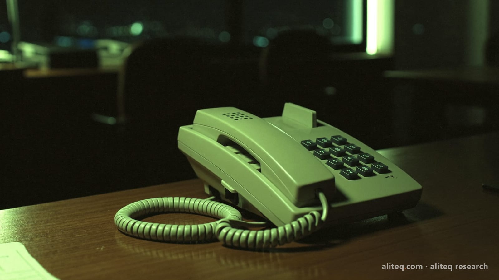 An old beige desk telephone with a coiled cord on a wooden desk in a dark office at night, lit by a faint lime-green glow