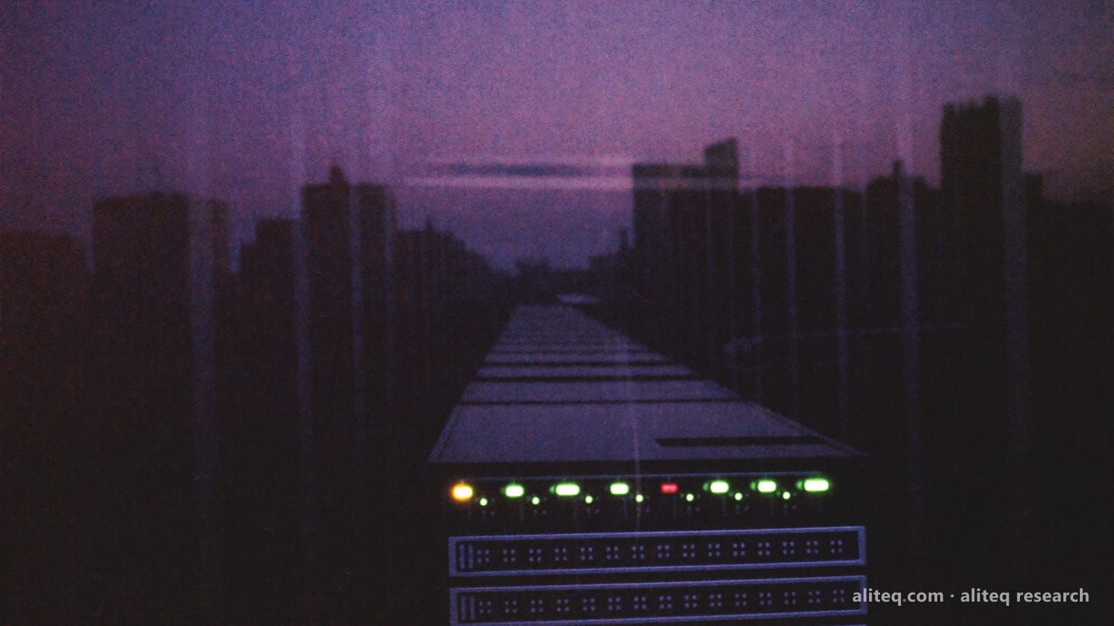 A dark server rack behind a glass wall at dusk, its row of green status lights glowing, with a violet city skyline reflected in the glass