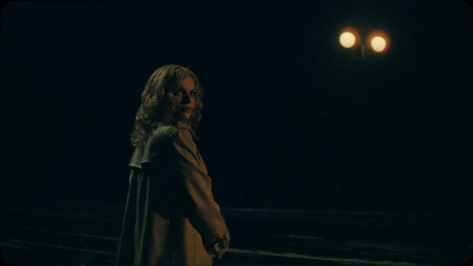 Cinematic night still of a fictional woman in a trench coat on an empty wet street, looking back over her shoulder, lit by two warm amber streetlights against deep shadow, with film grain and halation, in a 1970s neo-noir look