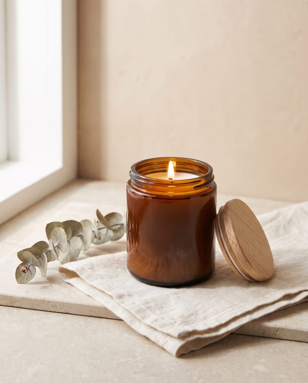 Product photo made with Higgsfield Marketing Studio Image on 26 October 2026: a plain unbranded soy candle in an amber glass jar with its wooden lid leaning beside it, lit wick, on a folded off-white linen cloth on a stone sill, with a sprig of dried eucalyptus and soft window light. The product is fictional; there is no label or text.