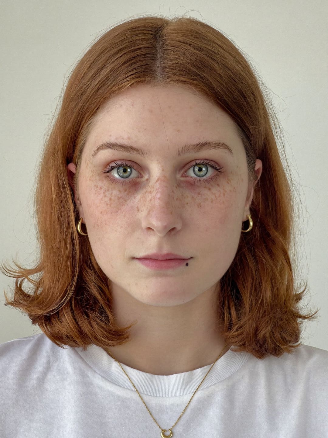 AI reference portrait of a fictional woman with grey-green eyes, a dense spray of freckles across her nose and cheeks, copper shoulder-length hair with a middle part, a small dark mole just below the right side of her lower lip as you look at her, small gold hoop earrings and a thin gold chain with a small crescent-moon pendant, in a white t-shirt on a grey background
