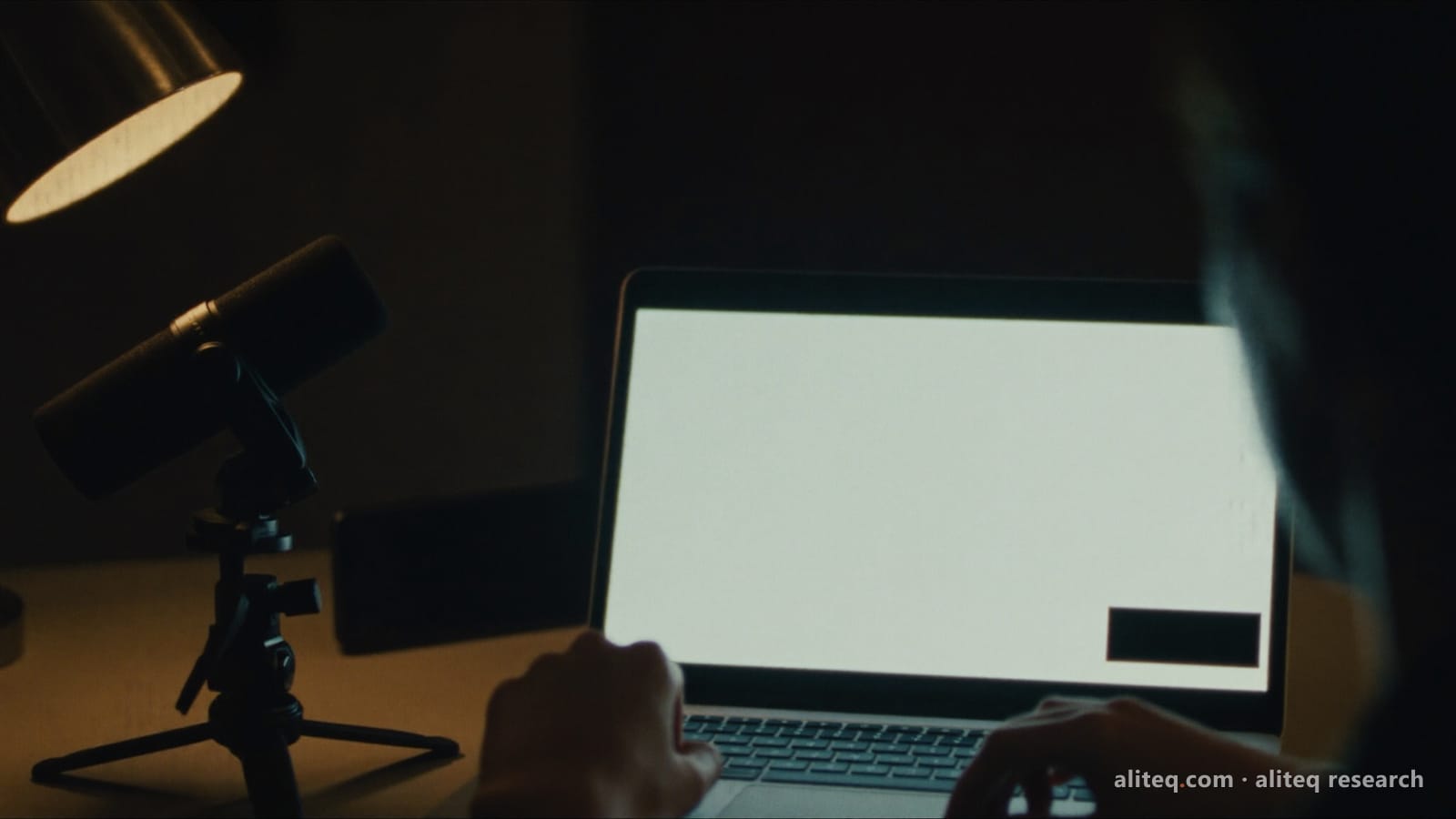 Photo of a creator at a dark desk at night, hands on a laptop whose blank white screen shows a small dark label box in the corner, beside a microphone on a tripod and a desk lamp