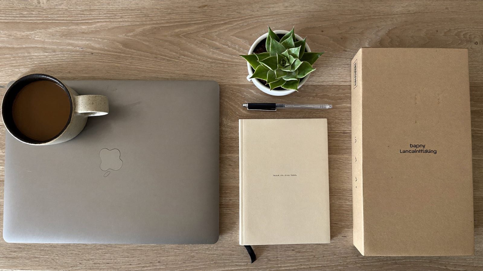 A top-down launch-day desk flat lay: a closed laptop, a coffee mug, a notebook, a pen and a plain product box