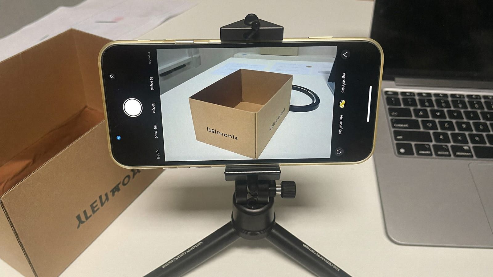 A phone on a small tripod capturing a plain product box next to a laptop on a bright home desk