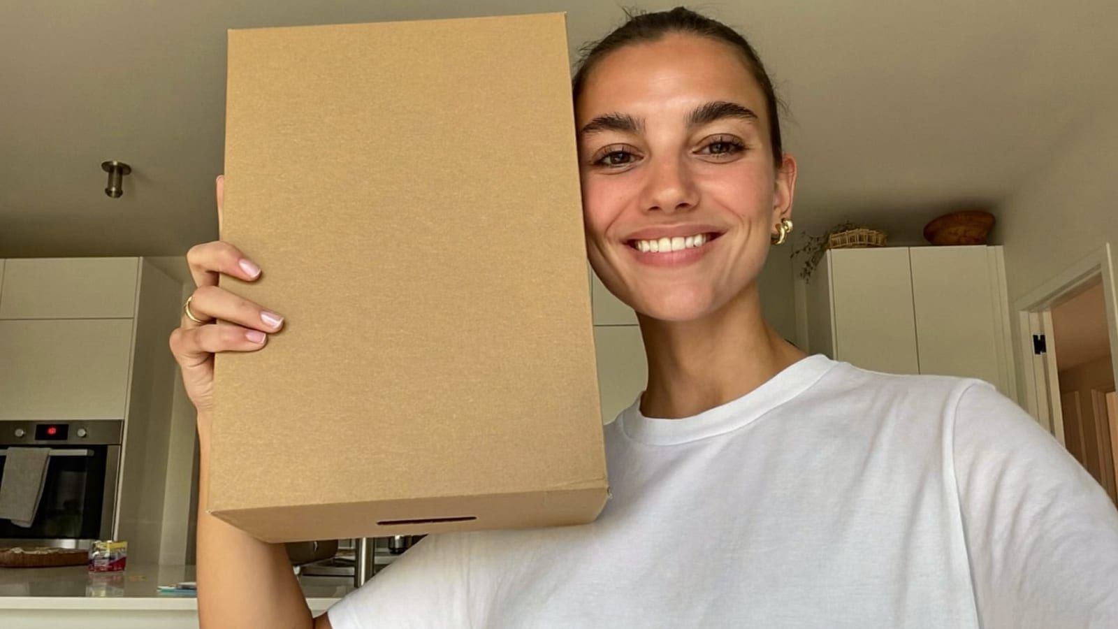 A UGC-style ad frame: a person holding a plain product box to the camera in a bright kitchen