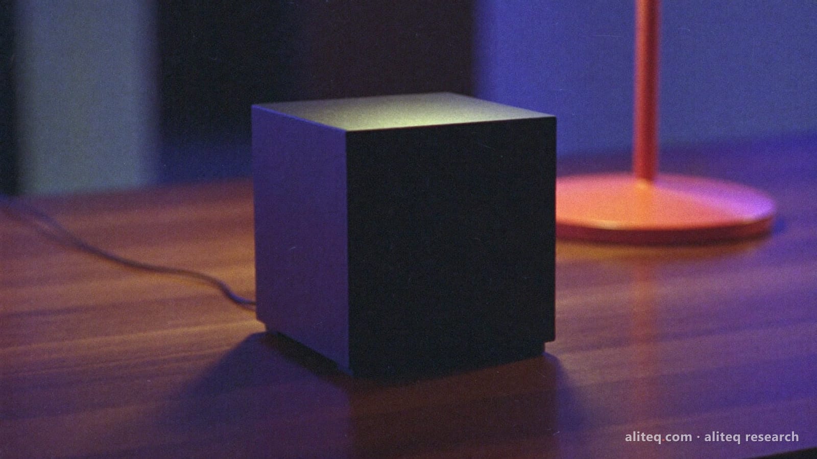 Photo of a small plain black cube computer on a dark wooden desk at night, a thin cable trailing off, indigo-violet light behind it and a coral desk lamp in soft focus