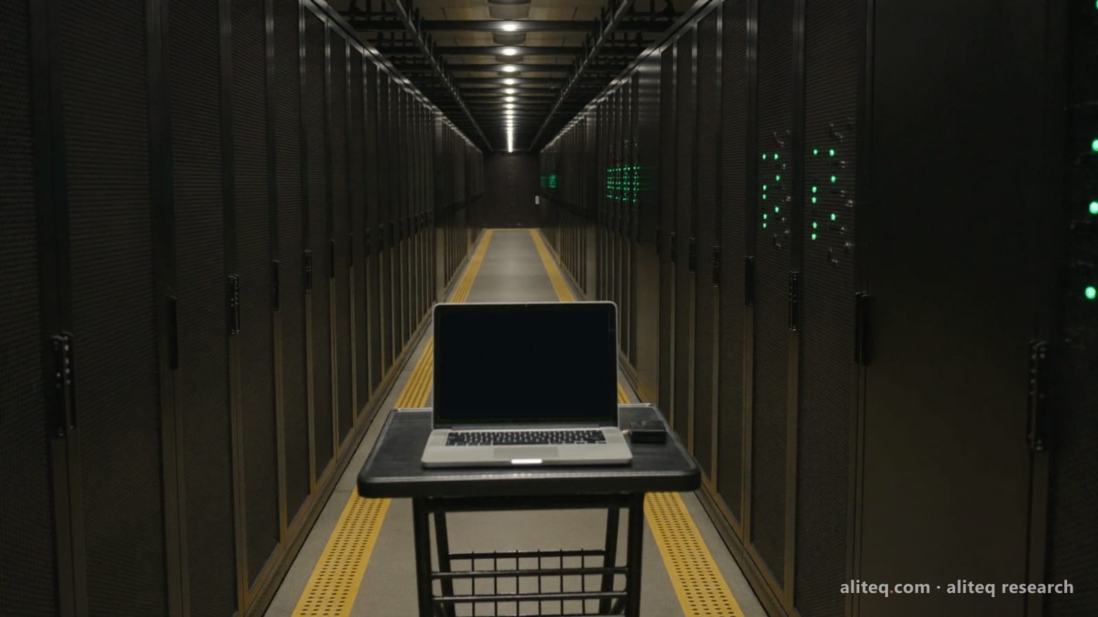 A laptop with a blank dark screen on a rolling cart in a long, dim data-center aisle lined with black server racks and a few green status lights