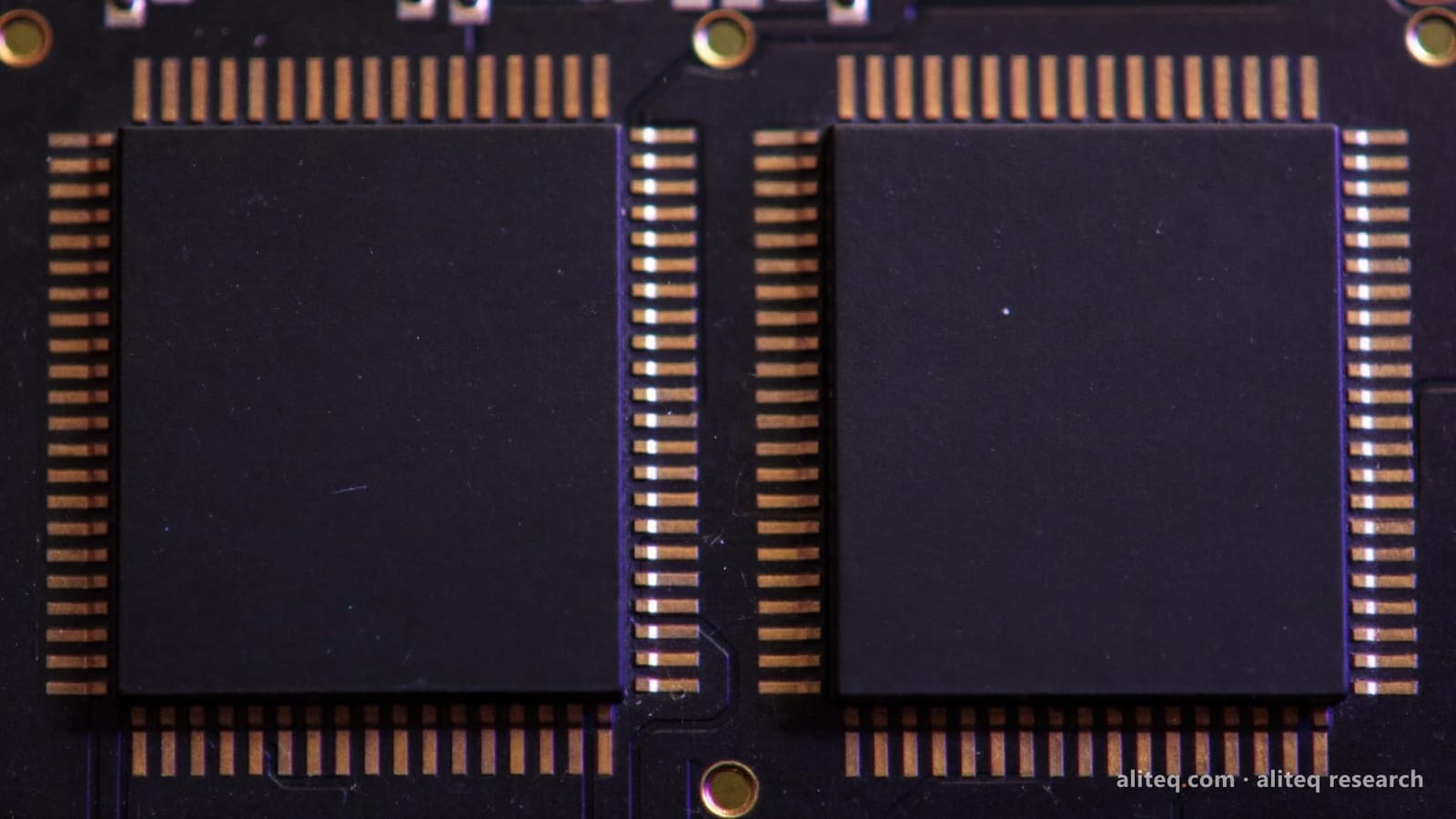 Macro photo of two identical blank black chips side by side on a dark circuit board with gold contact pads, lit in indigo-violet light