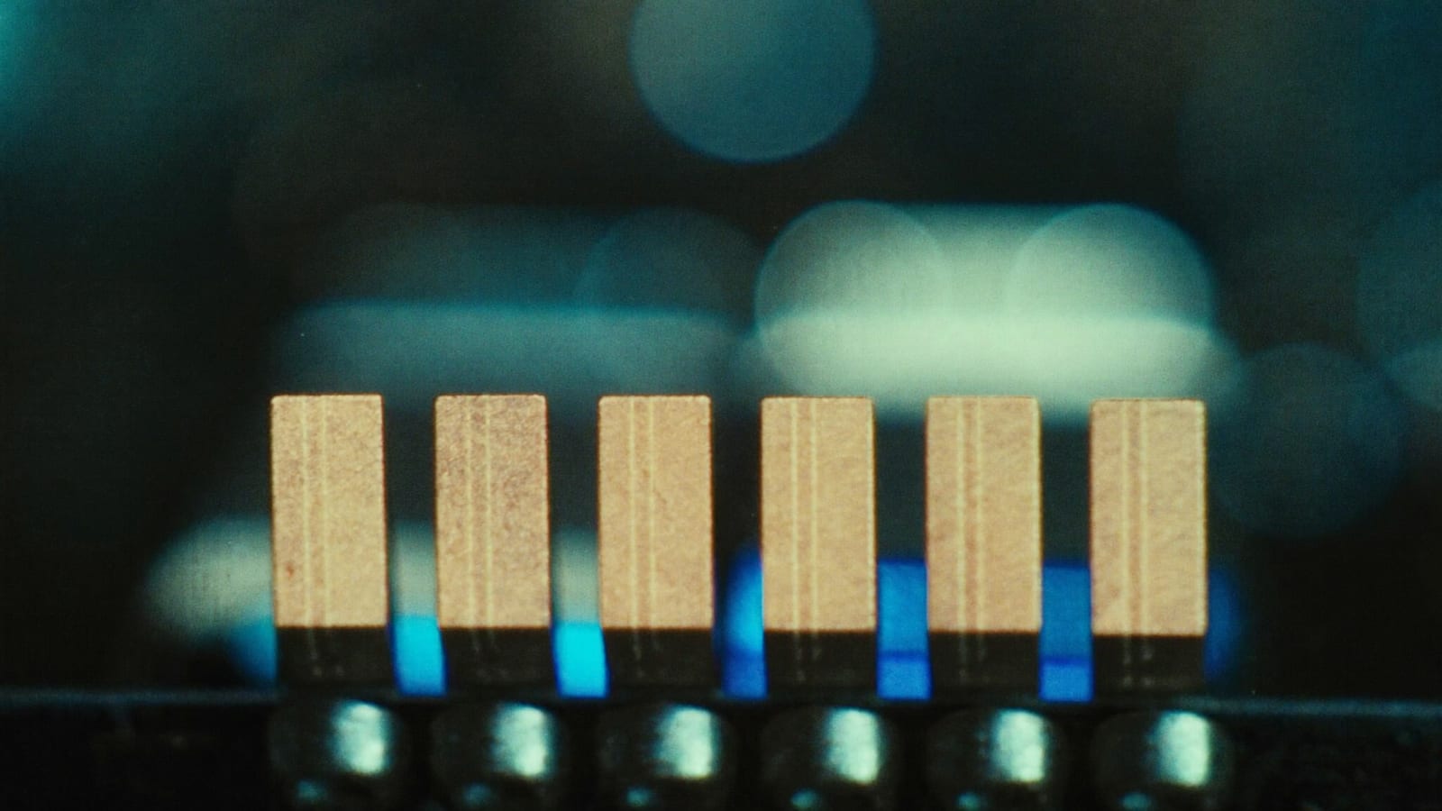 Extreme macro photograph of the gold contact fingers of a computer memory module, lit with cool blue accent light against a dark, softly blurred background