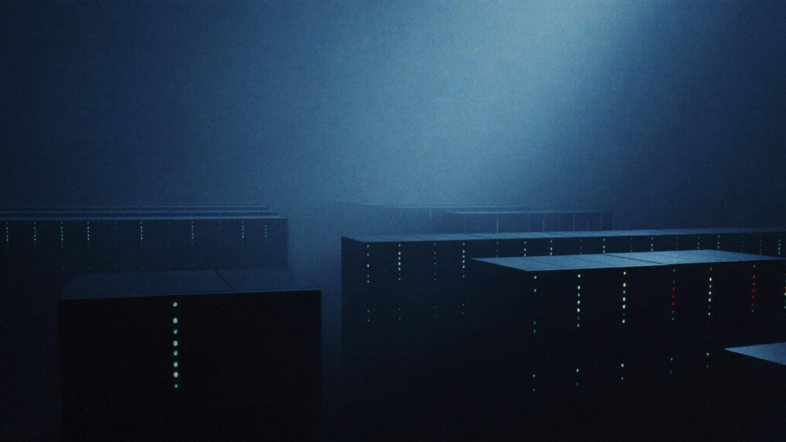 Photographic wide shot of a dark, hazy data-center hall, rows of server racks lit only by small green and red indicator lights, conveying large scale