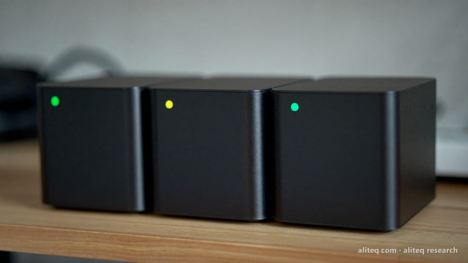 Three identical black compact servers side by side on a wooden shelf, two with green status lights and the middle one with a yellow light