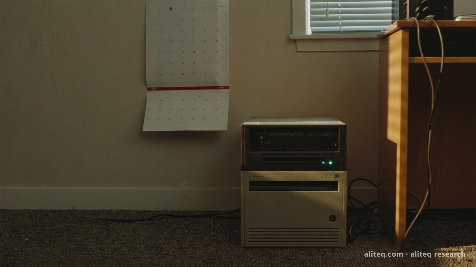 An old beige tower server on the carpet beside an office desk in late afternoon light, with a paper wall calendar pinned on the wall above it
