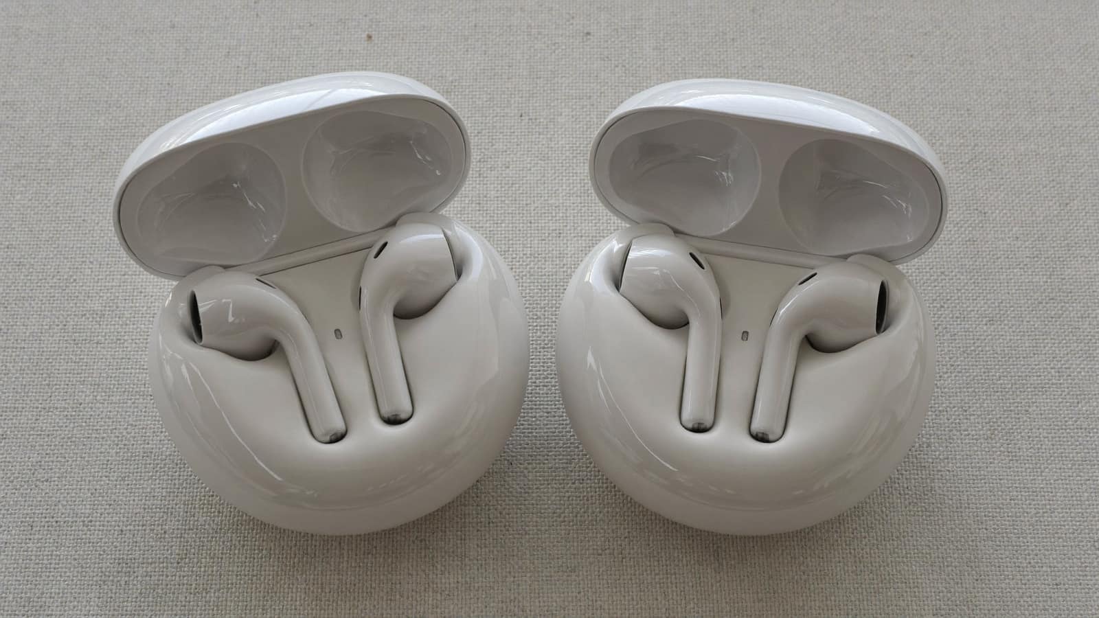 Two pairs of white wireless earbuds in open charging cases side by side