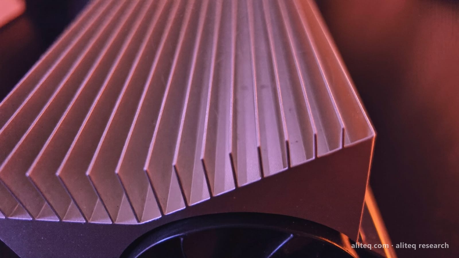 Macro photo of the silver aluminum heatsink fins of a graphics card above a black fan, lit in violet and warm coral light
