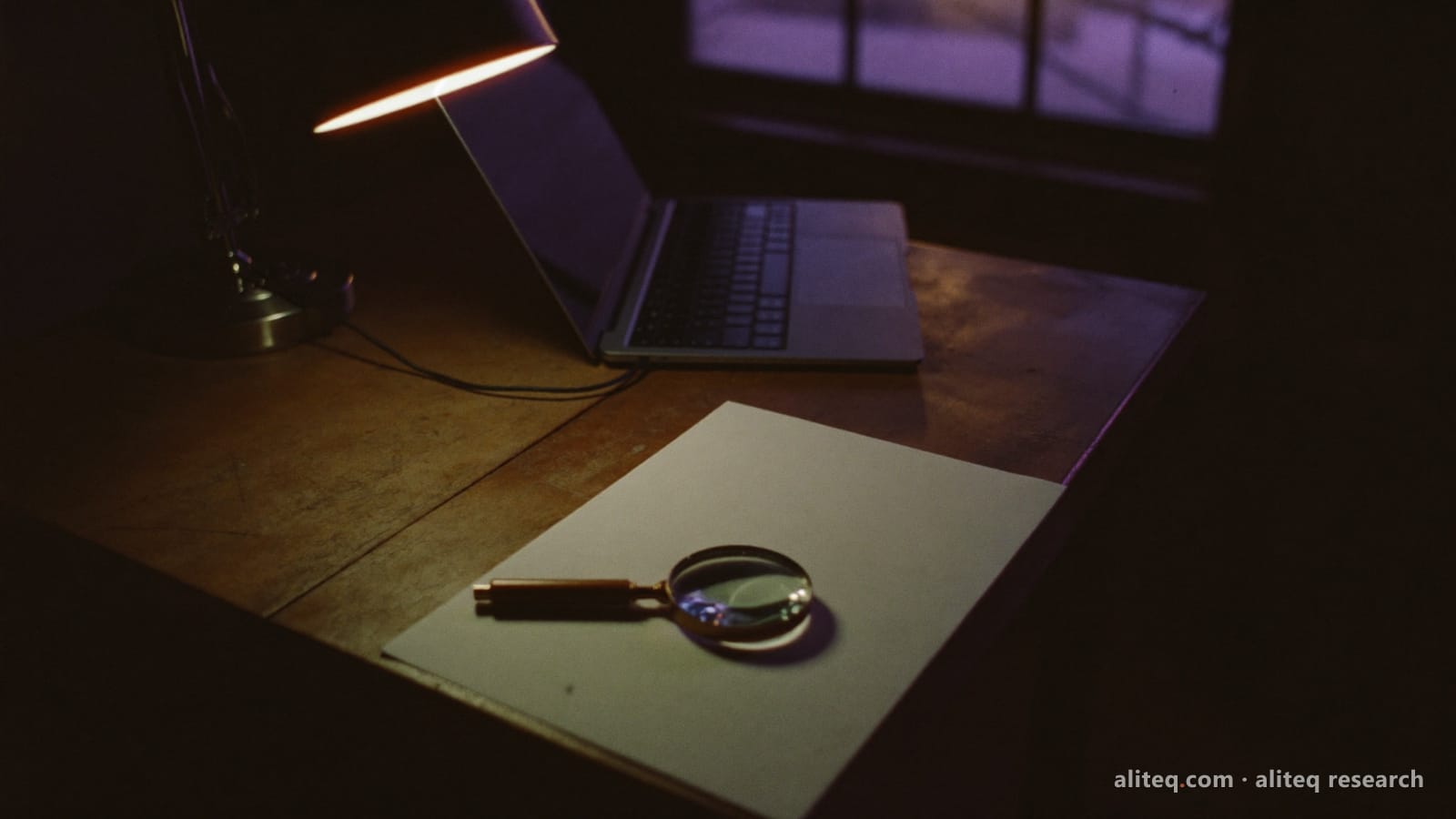 Photograph of a dark wooden desk at night, a desk lamp lighting a blank sheet of paper with a magnifying glass on it, an open laptop beside it and violet light through the window